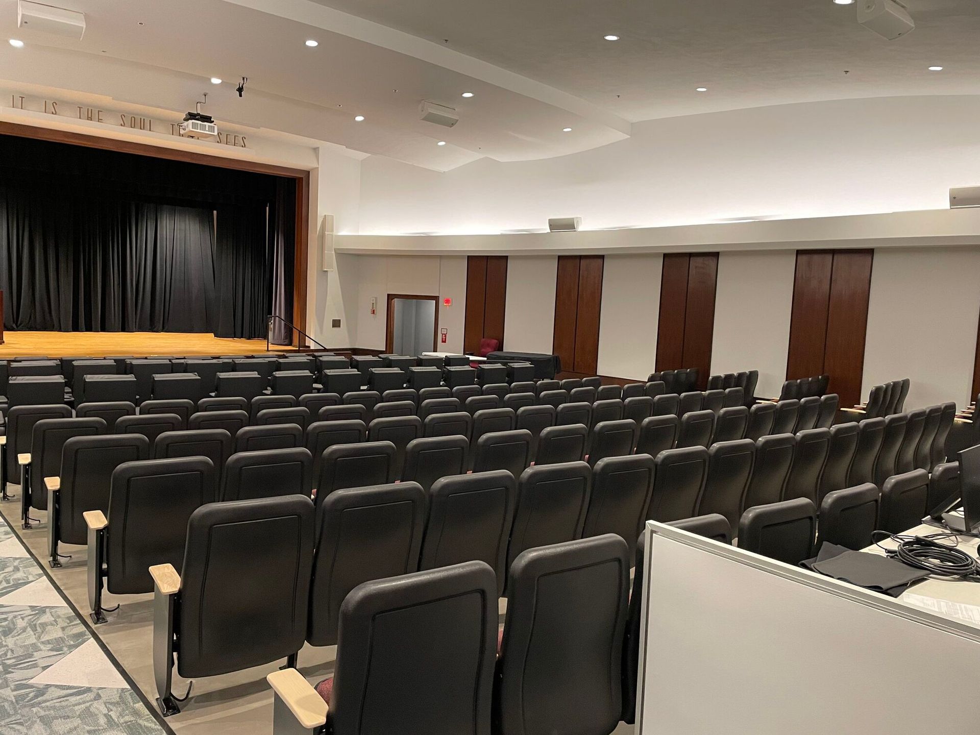 An empty auditorium with rows of black theater seats facing a stage with dark curtains.