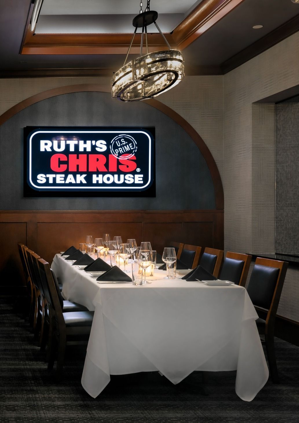 A formal dining room featuring a long table with white linens and place settings below a glowing Ruth’s Chris logo sign.