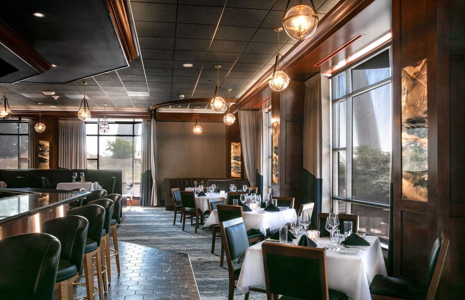 Upscale restaurant interior with dark wood, green upholstered chairs, white-clothed tables, and warm globe lighting.
