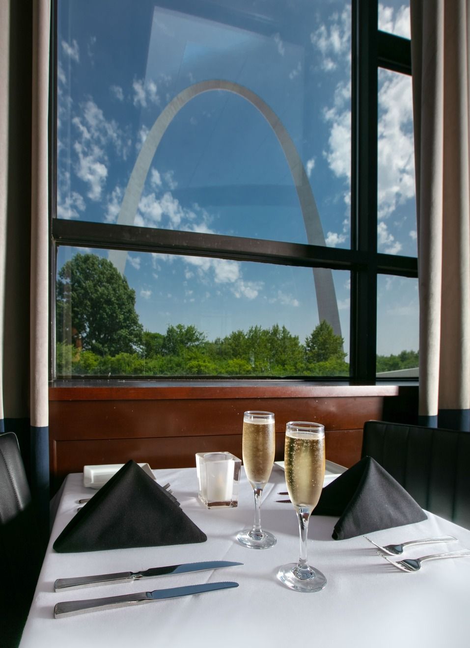 A table set with two champagne flutes and black napkins, overlooking the Gateway Arch in St. Louis through a window.