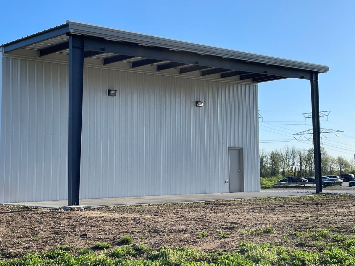 A white, metal-sided open-air pavilion with dark steel beams and support columns stands on a dirt lot under a blue sky.