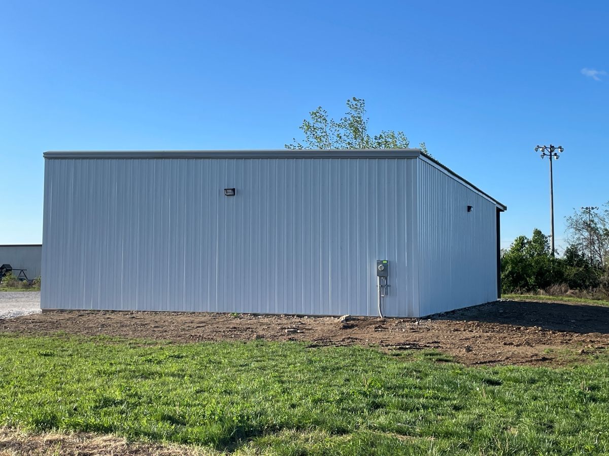 White metal warehouse building under a clear blue sky, set on a plot of grass and dirt.