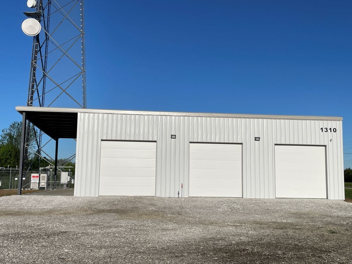 A white metal building with three garage doors and a tall communication tower under a clear blue sky.