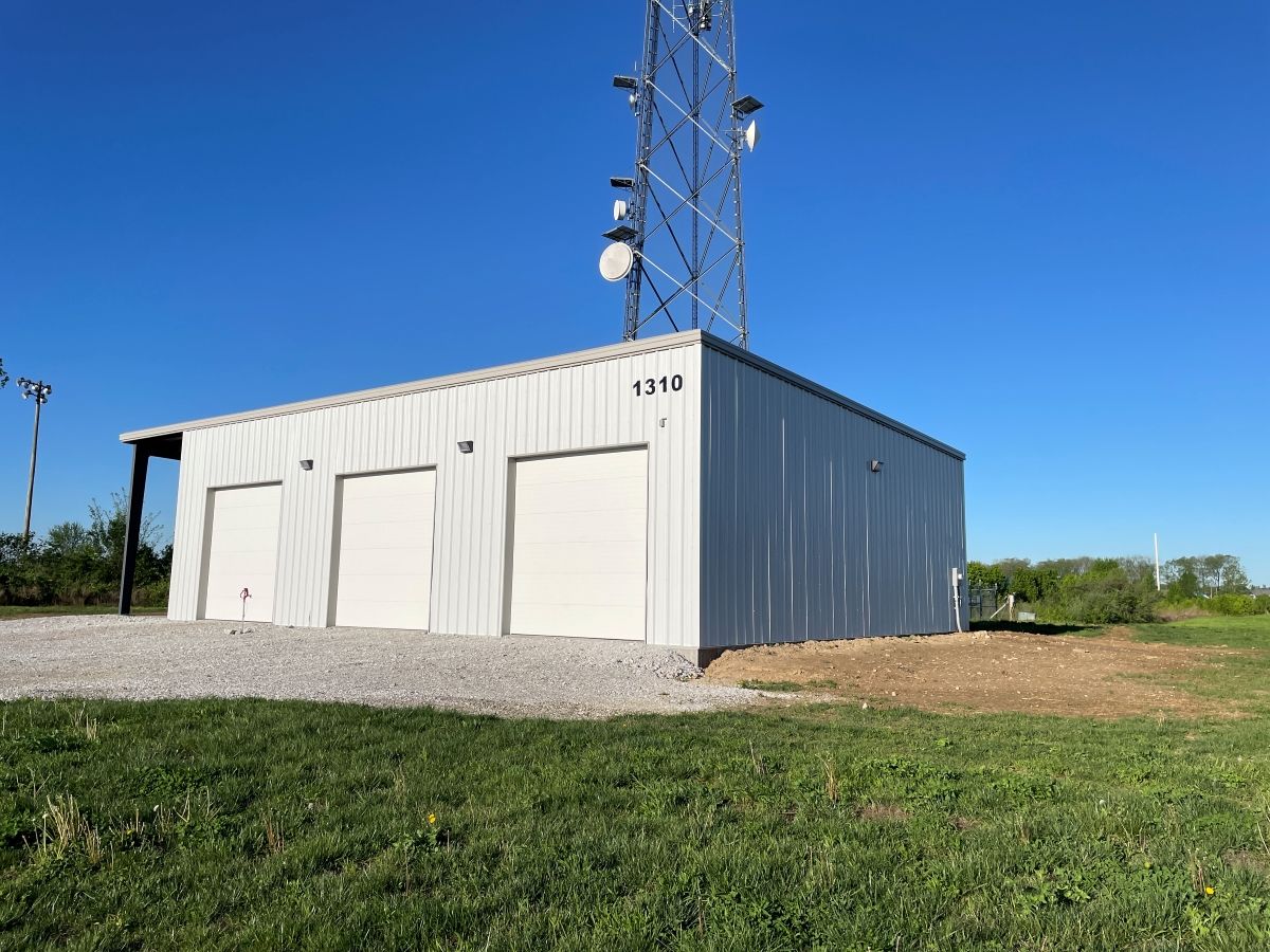 A metal storage building with three white garage doors stands next to a tall telecommunications tower under a blue sky.