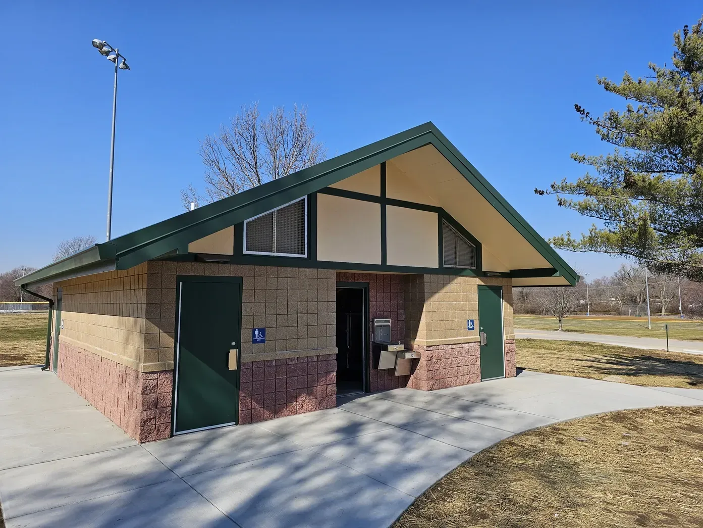 A public restroom building with stone-textured base, cream walls, dark green trim, and a gabled roof in a sunny park.