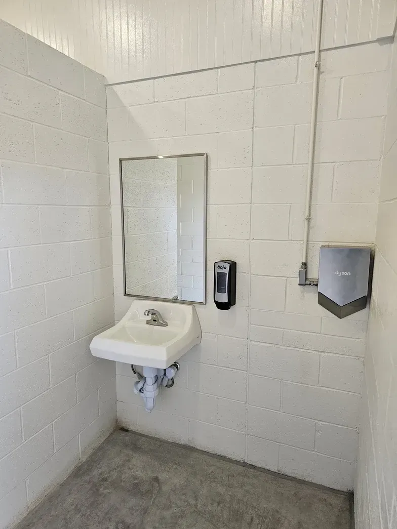 A white, wall-mounted sink with a mirror, soap dispenser, and hand dryer in a room with white cinder block walls.