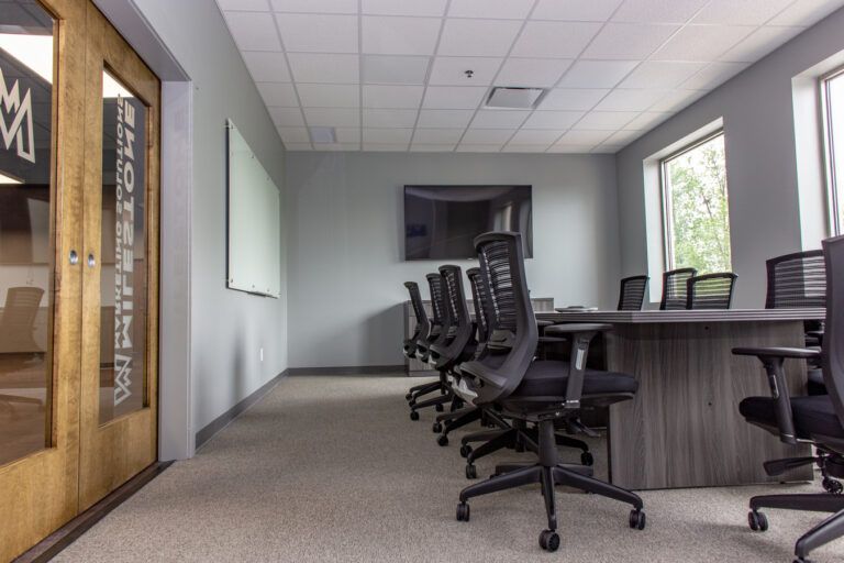 A modern conference room with a long table, ergonomic rolling chairs, a wall-mounted display, and large windows.