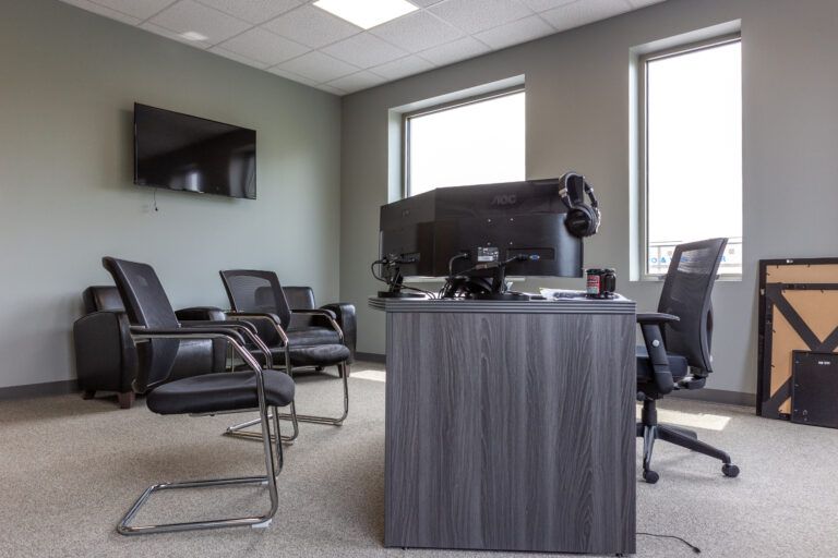 An office interior with a gray wooden desk, three black mesh chairs, a wall-mounted TV, and two windows.
