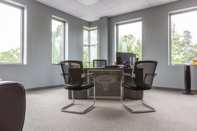An office with a desk featuring a Ford logo, two mesh chairs, and windows with a green view against light grey walls.