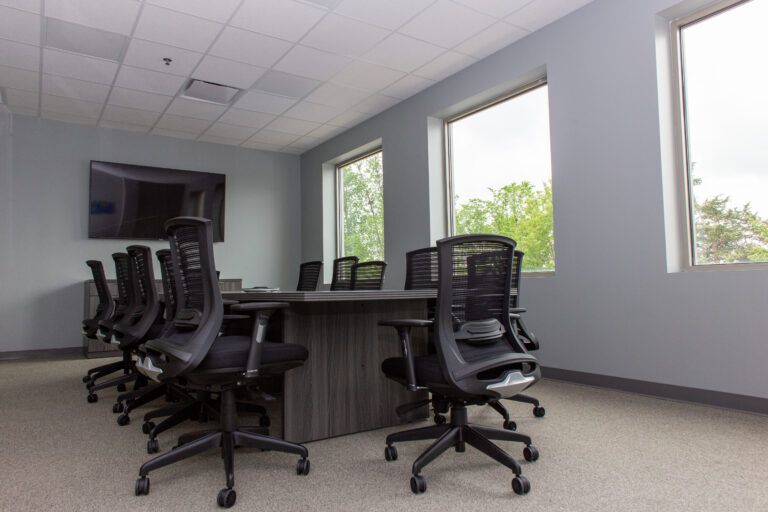 A conference room featuring a dark wooden table surrounded by several black ergonomic office chairs against gray walls.