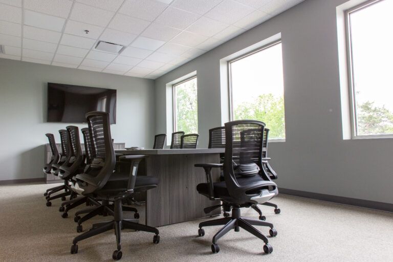 A modern conference room with a large rectangular table, mesh office chairs, and a wall-mounted flat-screen television.