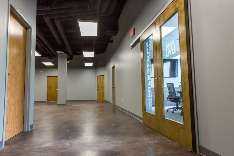 A hallway with wood-paneled doors, concrete floors, exposed pipes on a dark ceiling, and a glass-paneled office door.