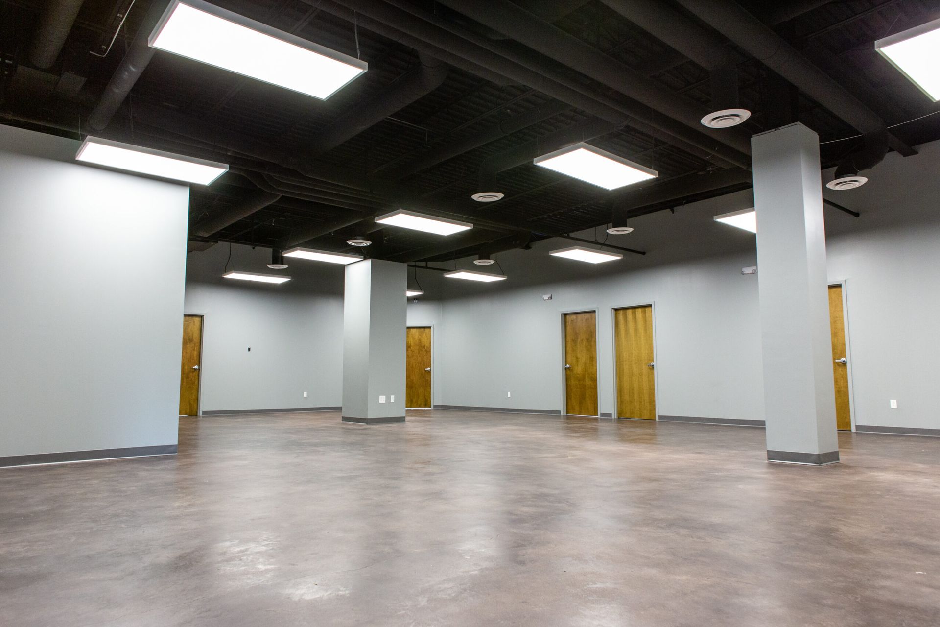 An empty office space with gray walls, concrete floors, a black open-plank ceiling, and several wooden doors.