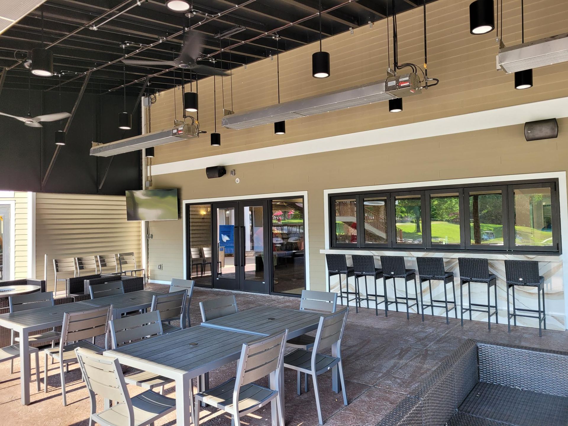 A covered outdoor patio with grey tables, chairs, and a bar counter with stools, featuring overhead heaters and lighting.