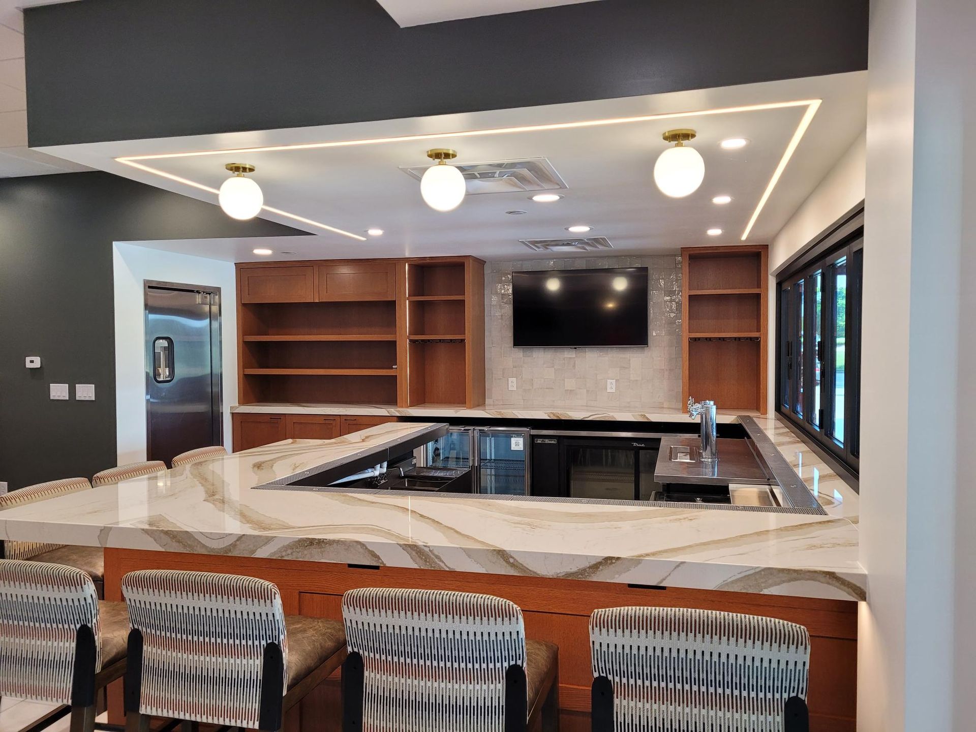 A modern indoor wet bar with a white stone countertop, wood cabinetry, four patterned barstools, and globe lighting.