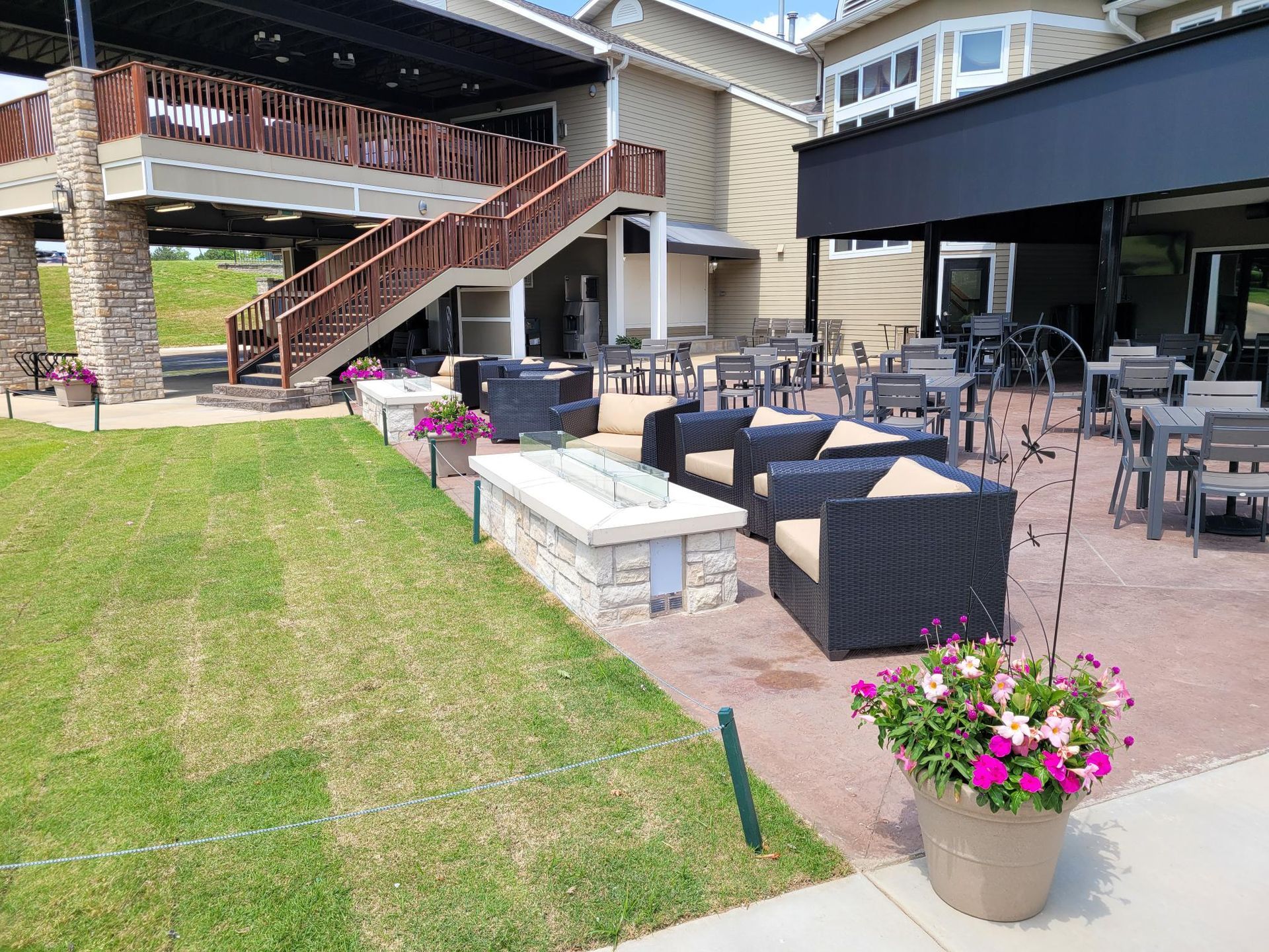 An outdoor patio with stone fire pits, wicker chairs, dining tables, a staircase, and potted flowers on a sunny day.