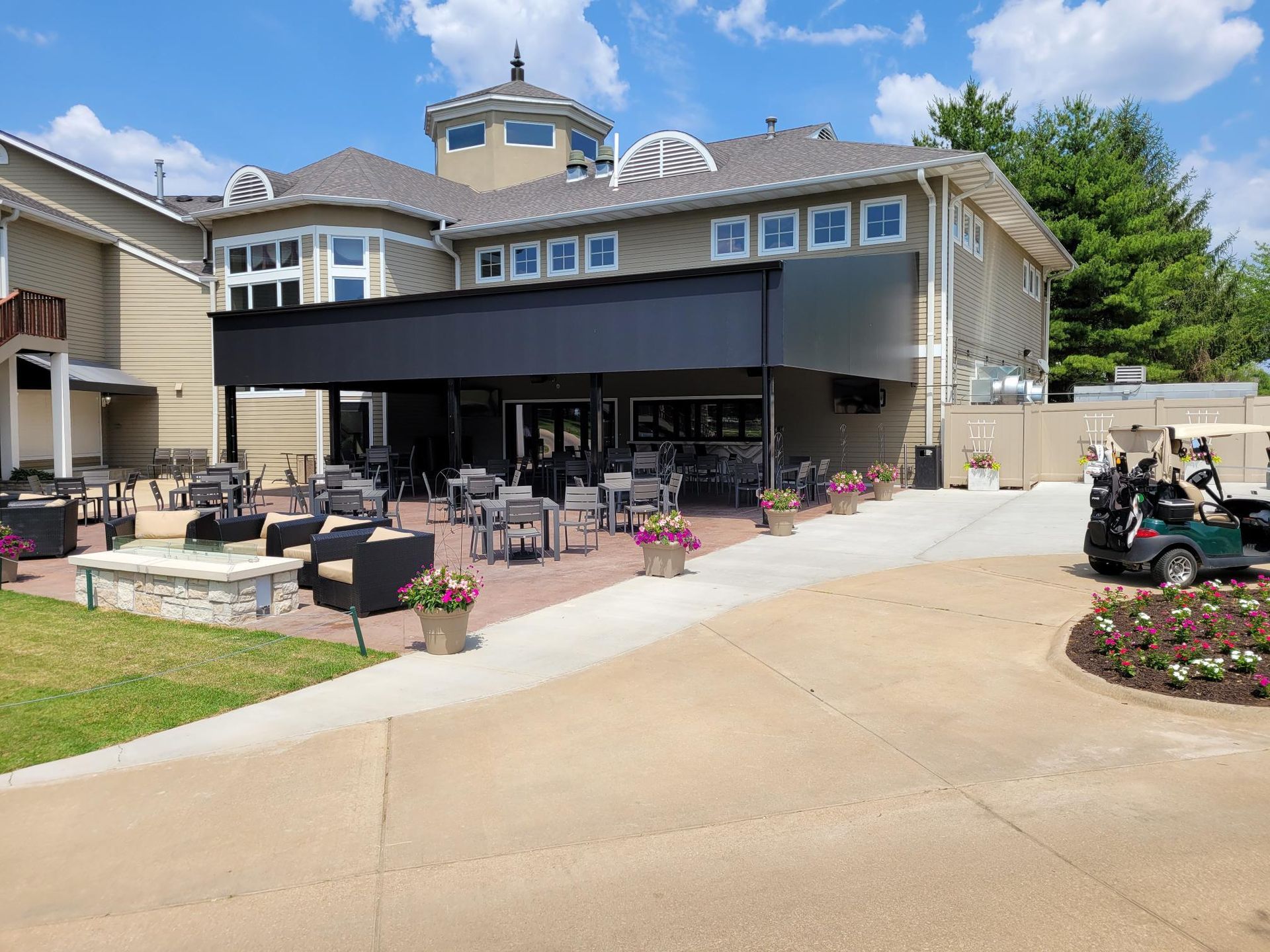 A sunny outdoor patio area at a multi-story building featuring tables, lounge chairs, a fire pit, and a golf cart.