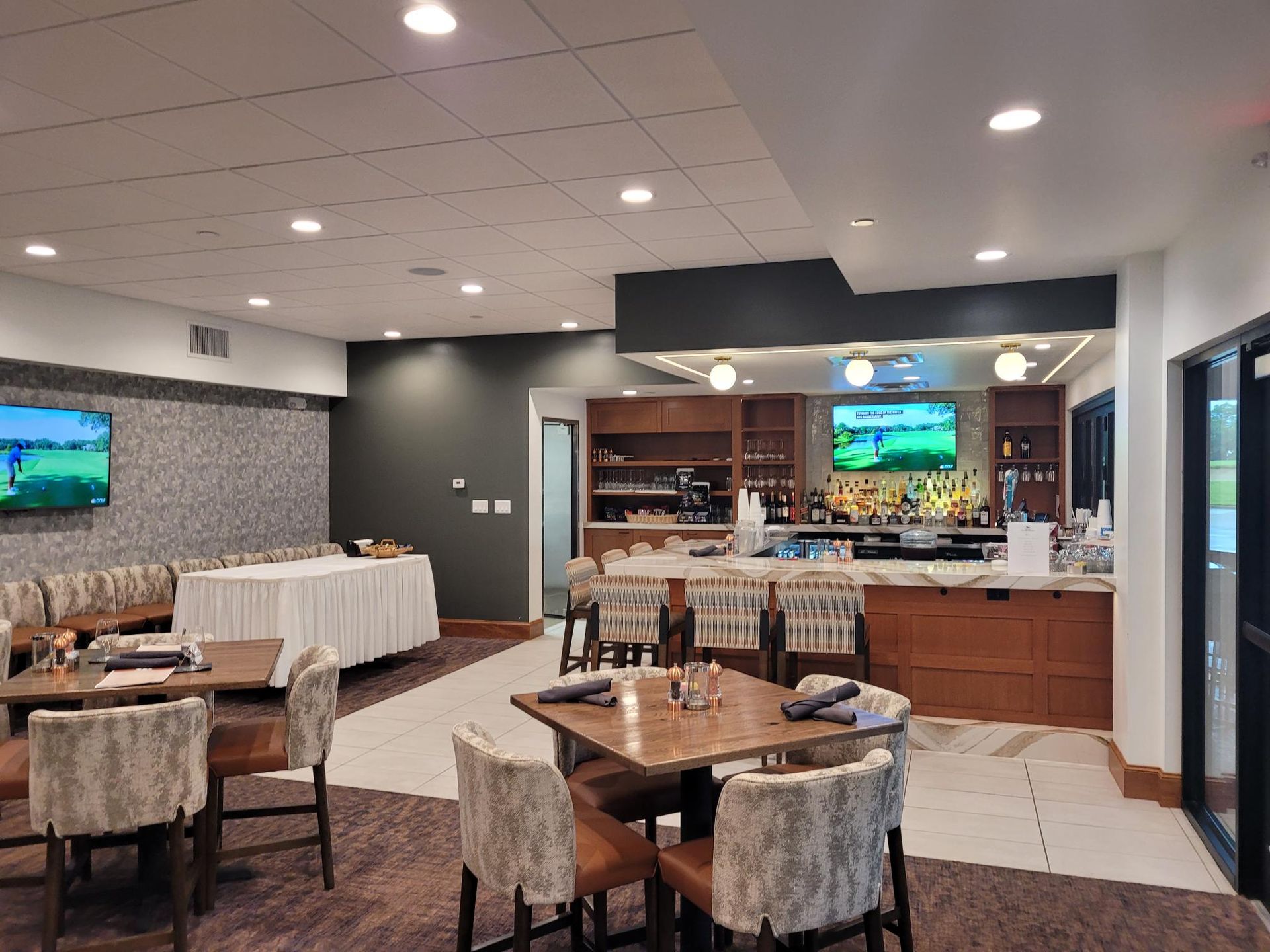 An indoor restaurant space with tables, patterned chairs, a long bar with stools, and televisions on the walls.