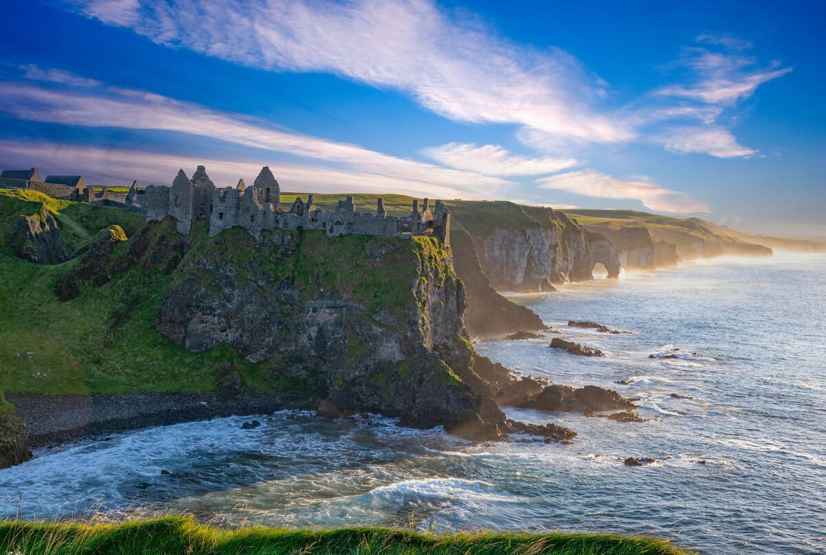 Ruins of Dunluce Castle on a green cliff overlooking the sea, under a blue sky with clouds.