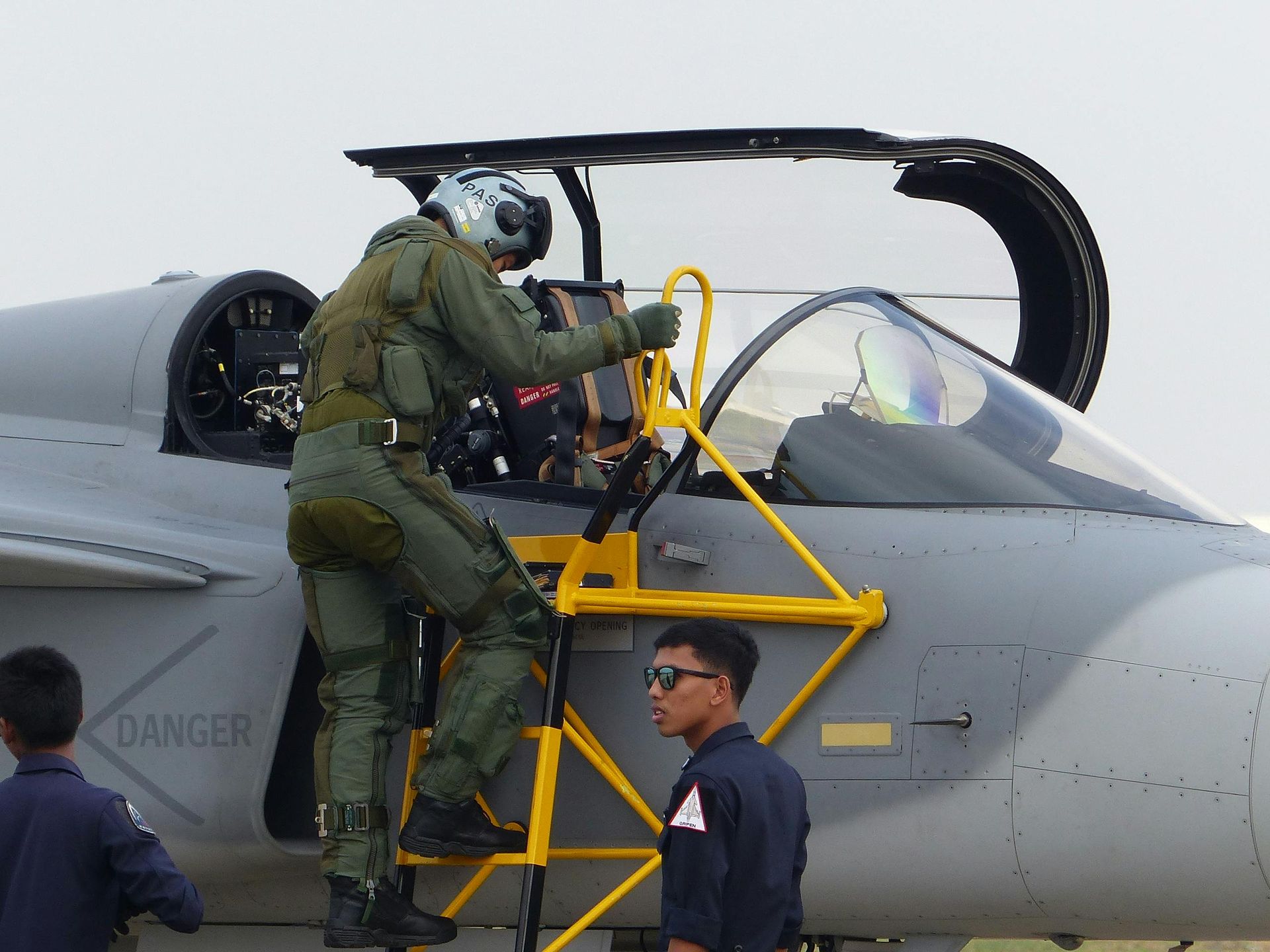 Pilot entering a gray jet cockpit via a yellow ladder, another person looks on.