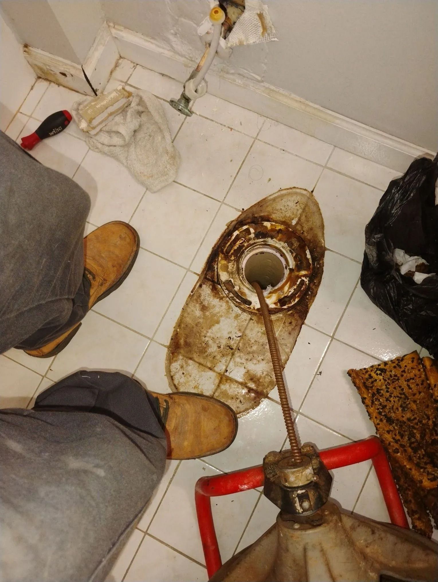 Worker inspecting a rusty floor drain in a dirty tiled utility room, with tools and boots visible.