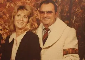 Two people posing together against an autumn-colored backdrop, both smiling and dressed formally.