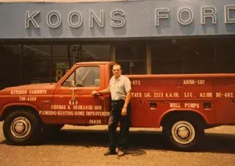 Man standing beside a red service truck in front of a storefront with “KOONS” signage.