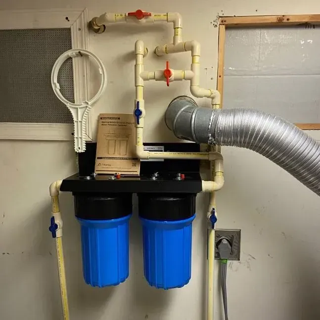Wall-mounted water filter system with two blue canisters, pipes, valves, and a silver vent hose in a utility room