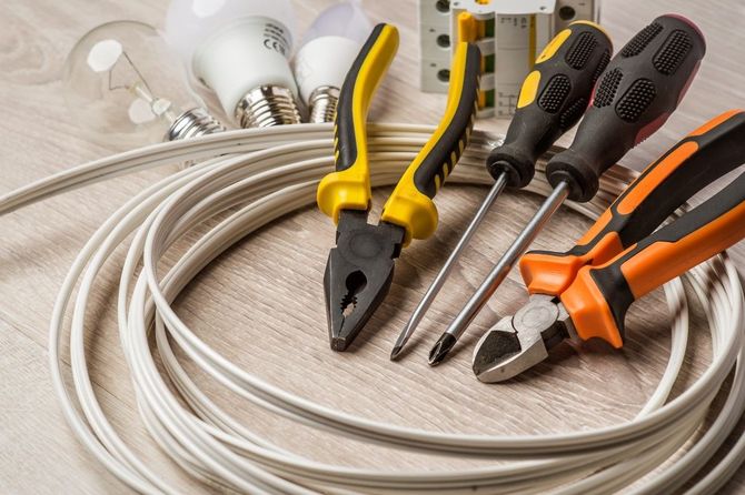 Assorted hand tools and coiled cables on a light wooden surface, including screwdrivers, pliers, and light bulbs