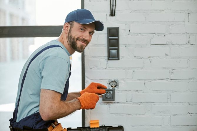 Handyman in blue cap and orange gloves using a tool on a wall outlet during home repair