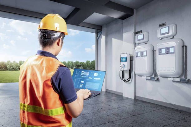 Worker in safety vest and hard hat using a tablet beside wall-mounted electrical boxes in a garage.