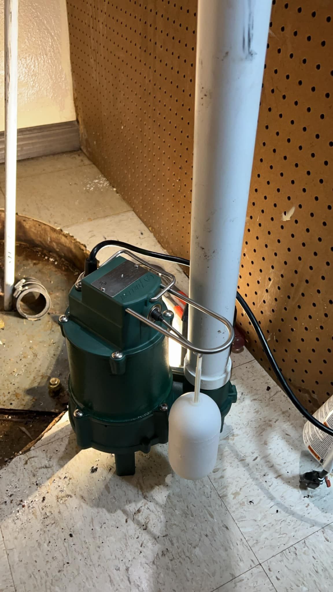 A green sump pump is sitting next to a white pipe in a basement.
