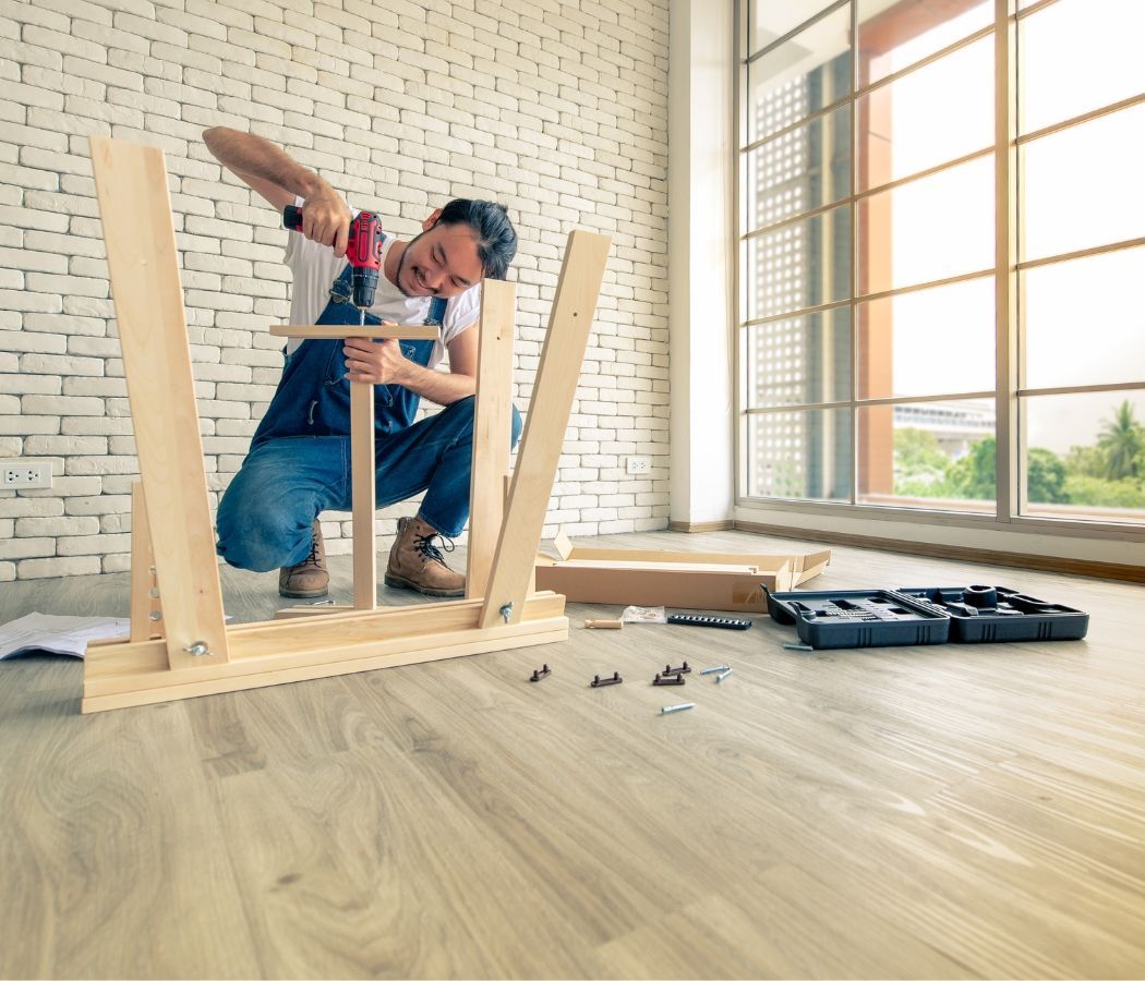 A man is kneeling down and using a drill to assemble a wooden table.