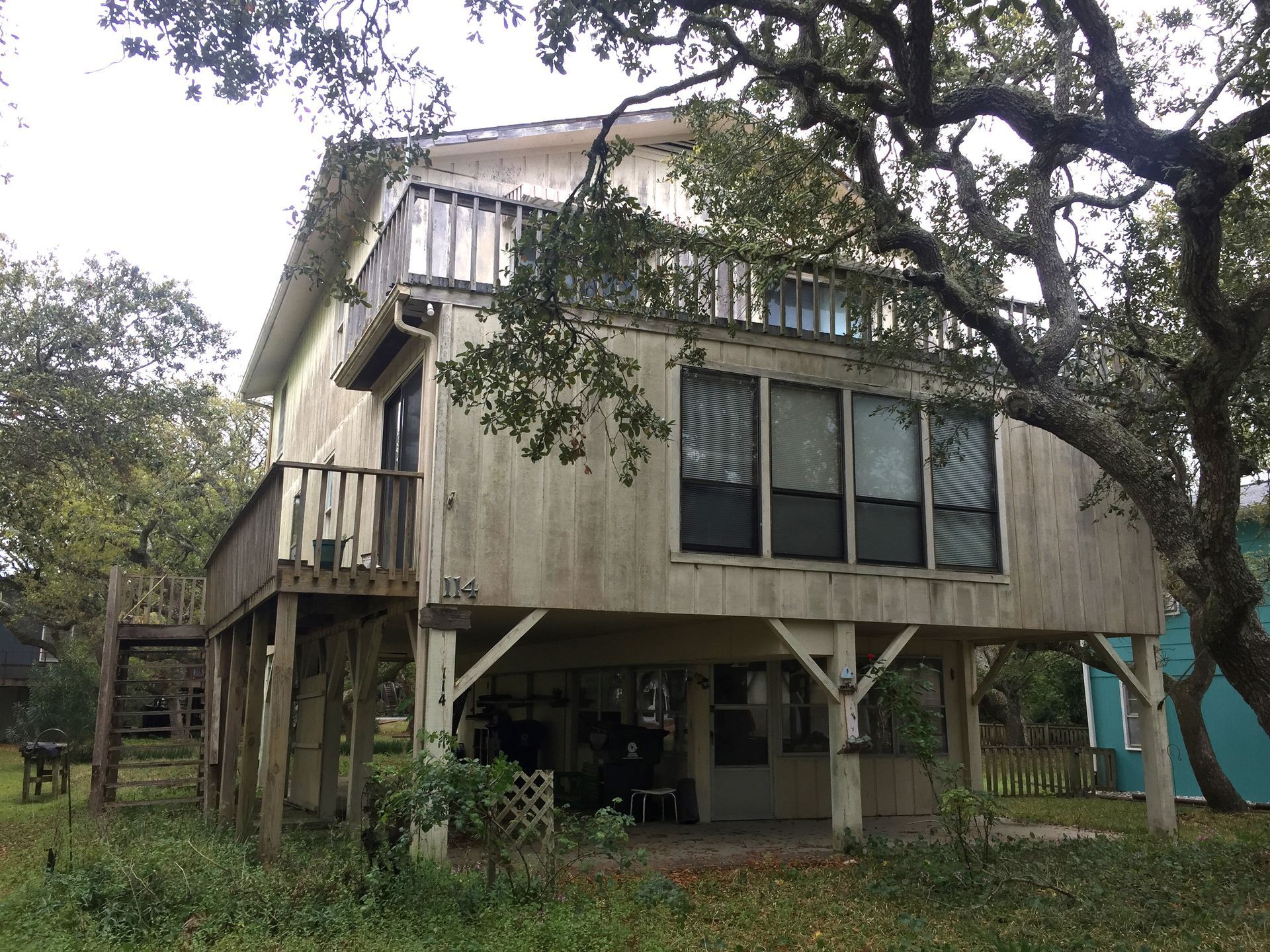 Two-story weathered beach house on stilts with balconies. Overgrown foliage surrounds the home.