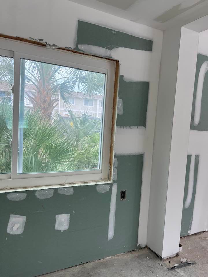 Interior room under construction, with green drywall, white trim, window, and mudding in progress.