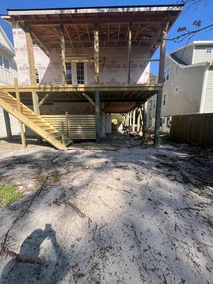 Two-story house under construction with a wooden deck and stairs, set in a sandy yard.