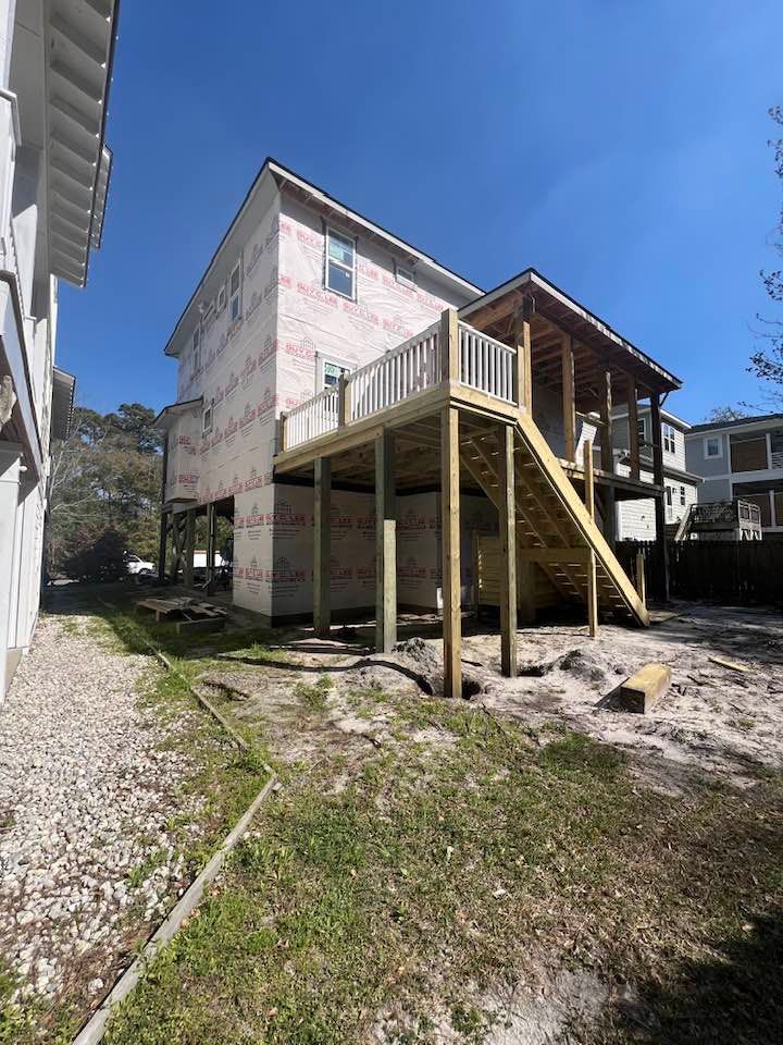 Two-story house under construction with a wooden deck and stairs, set on a sandy lot under a blue sky.