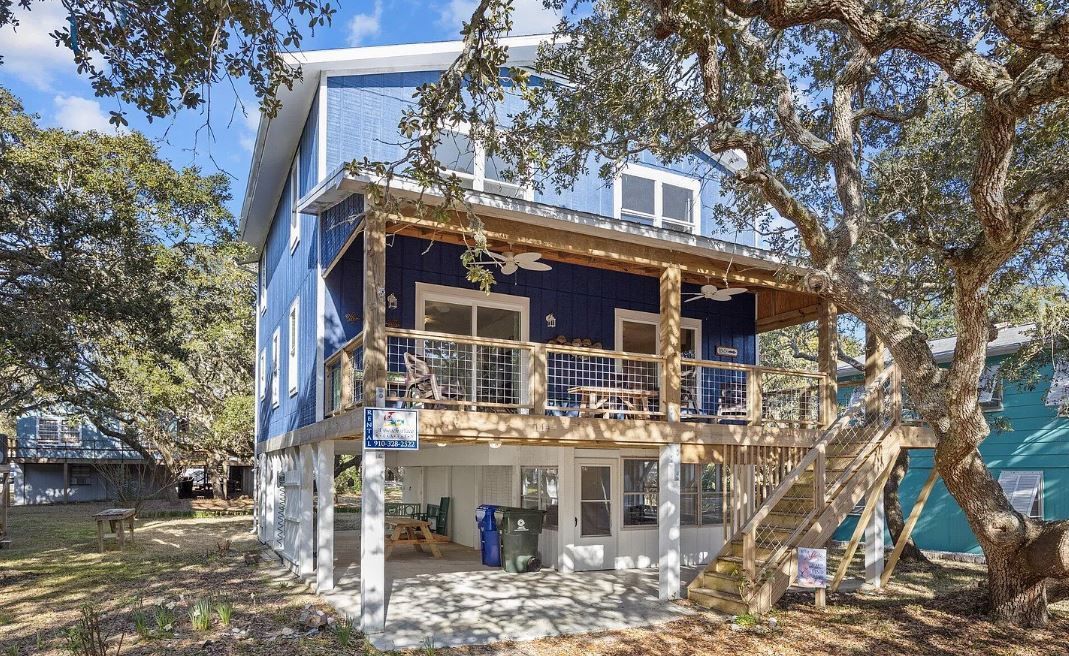 Blue two-story beach house with porch, white supports, and large tree in the yard.