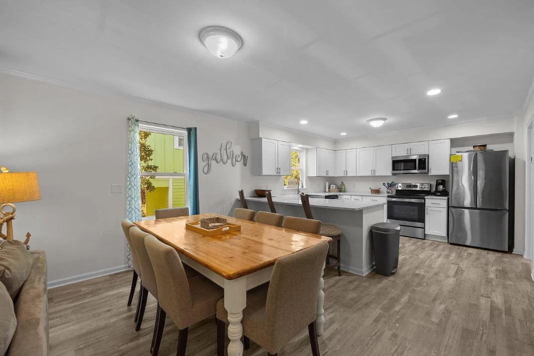 Dining room with wooden table, chairs, and kitchen with white cabinets, appliances.