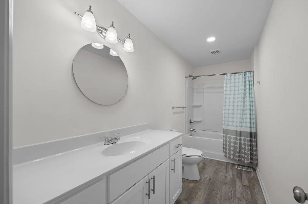 Bathroom with white vanity, round mirror, and shower with patterned curtain. Gray wood-look floor.