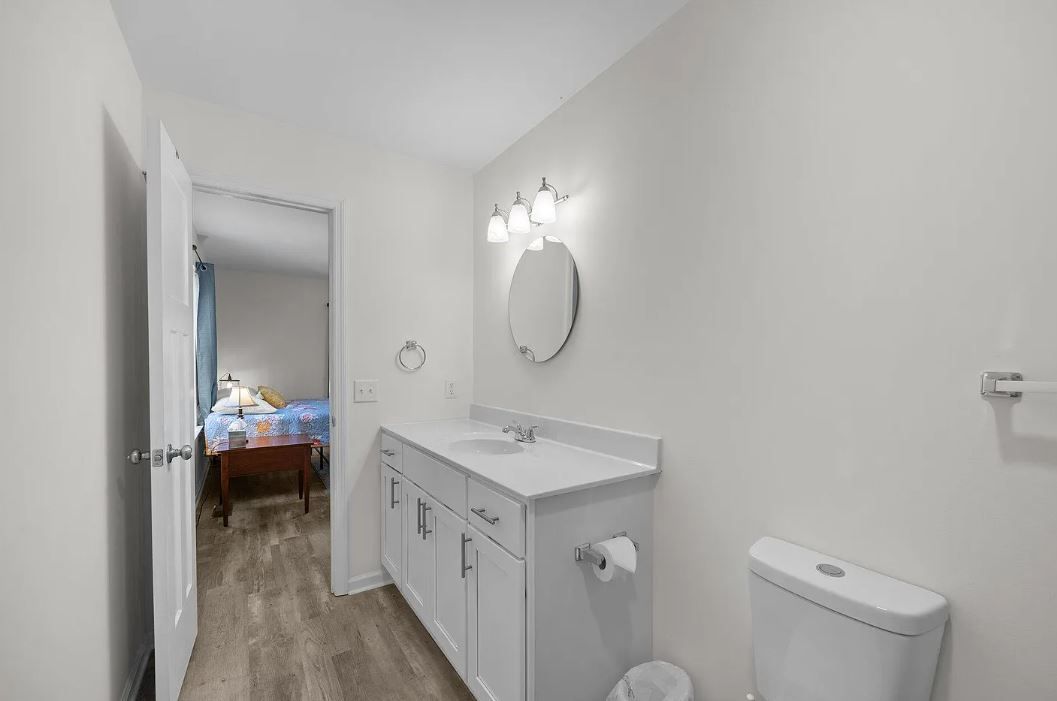 Bathroom with white vanity, mirror, toilet, and open doorway to a bedroom with bed.
