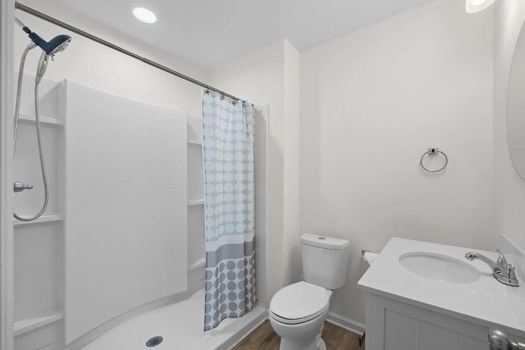 A white bathroom with a shower, toilet, and vanity. A blue shower curtain hangs, and the floor is wood-toned.