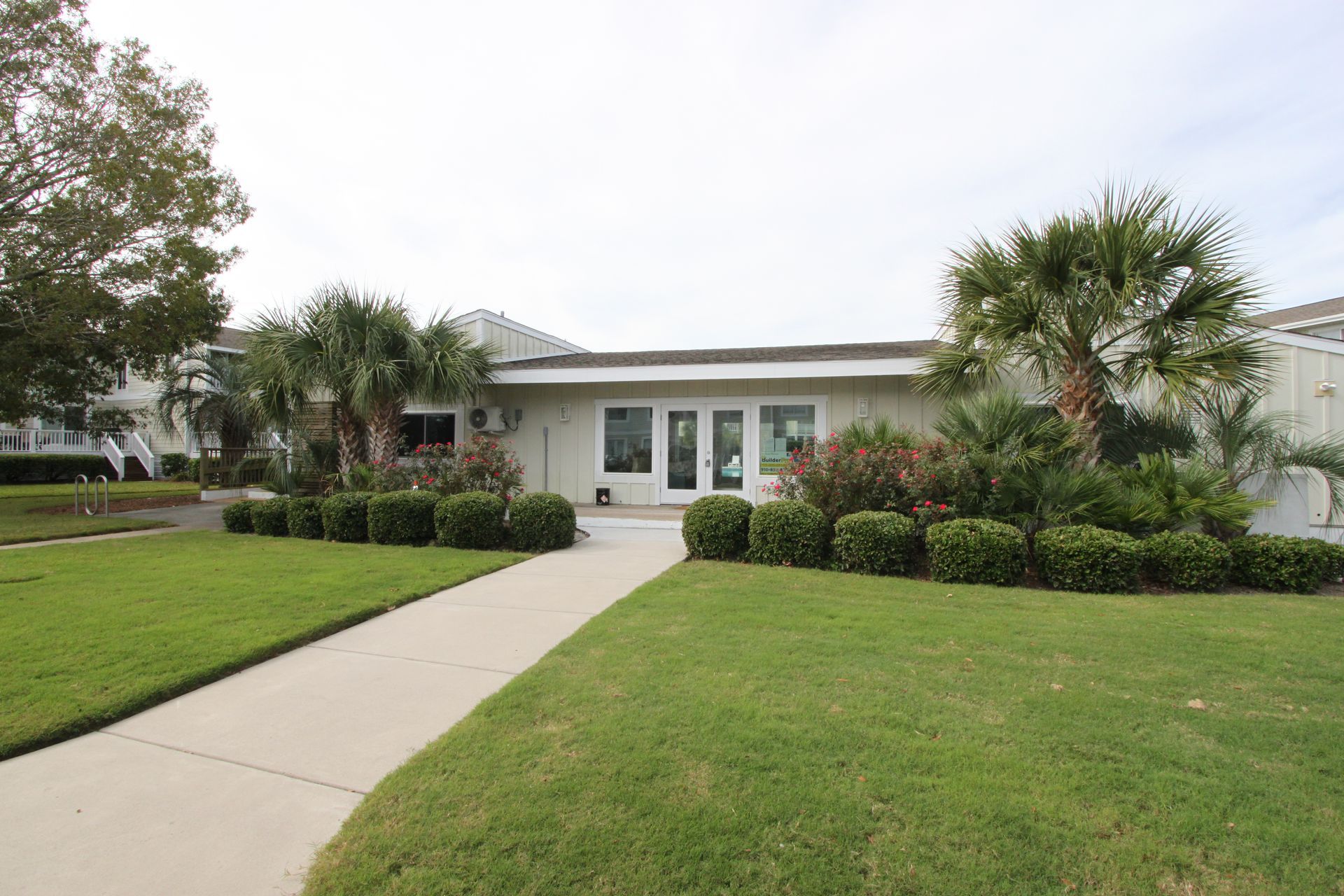 A one-story building with a walkway, surrounded by grass, bushes, and palm trees.