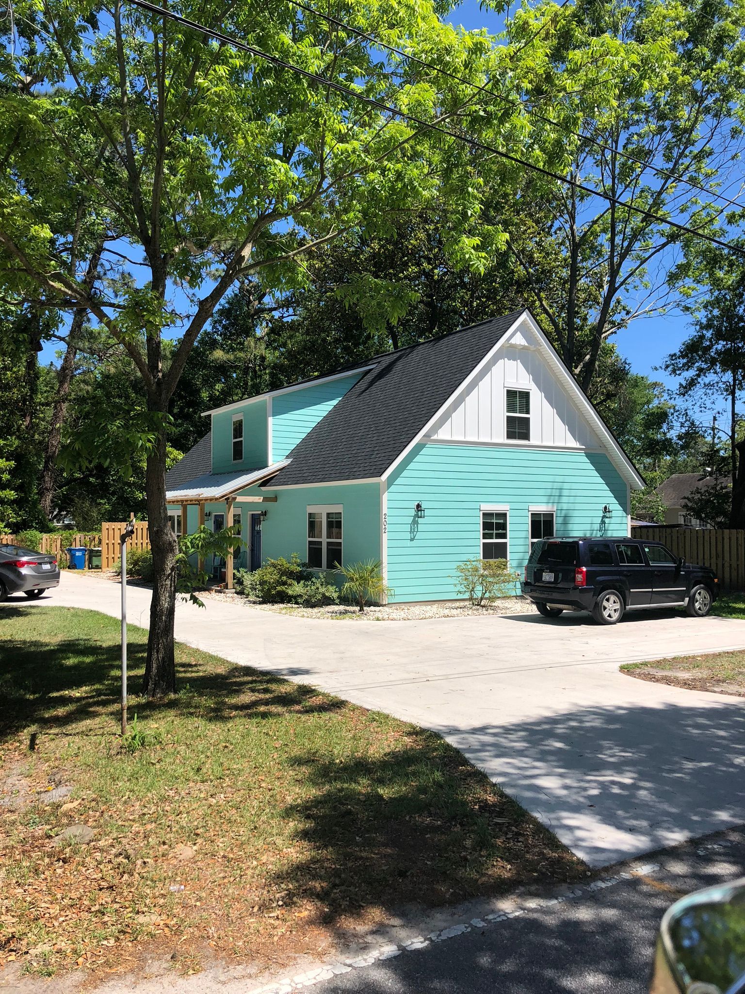 A turquoise house with a black roof and white trim, surrounded by trees, parked cars in a driveway.