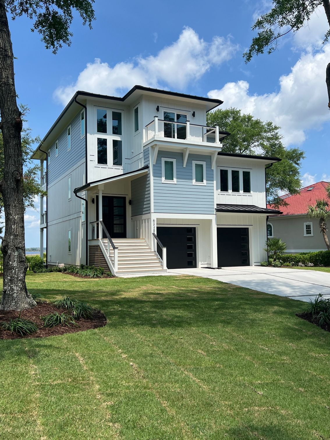 Three-story coastal home with light blue siding, white trim, black garage doors, and a balcony.