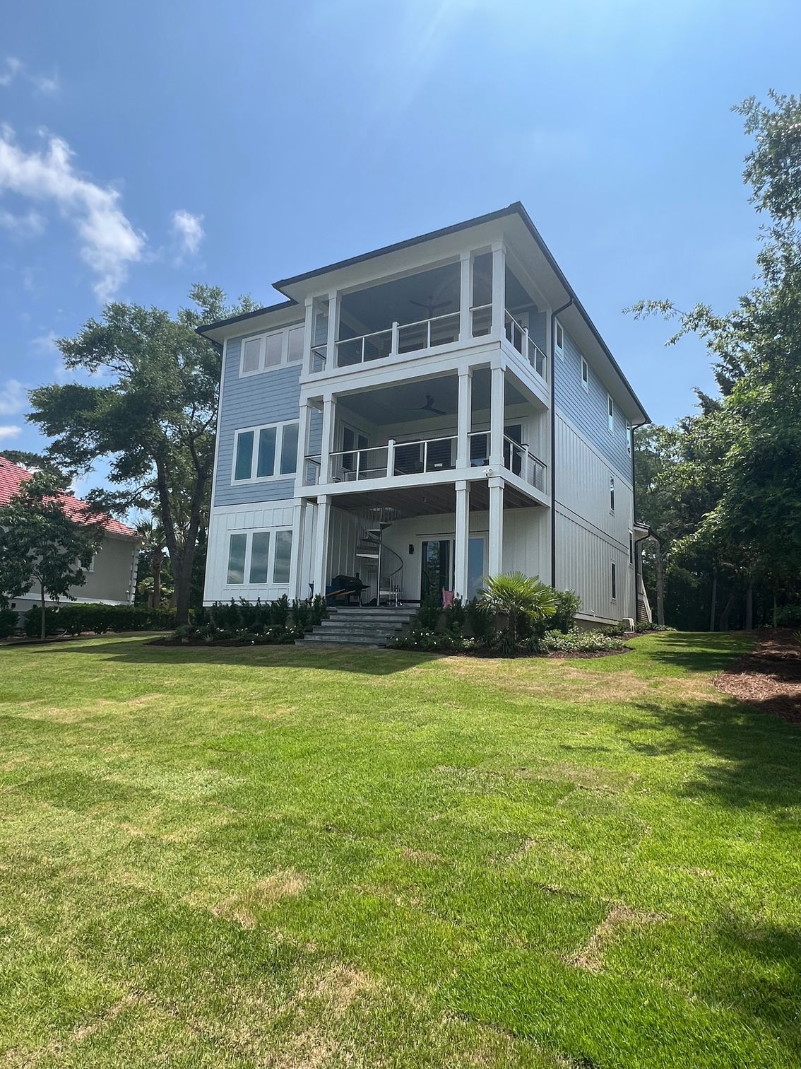 Three-story house with blue siding, balconies, white columns, and lush green lawn under a bright blue sky.