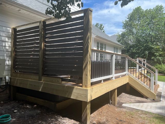 Wooden deck with a privacy screen next to a house with white siding, surrounded by trees and a paved area.
