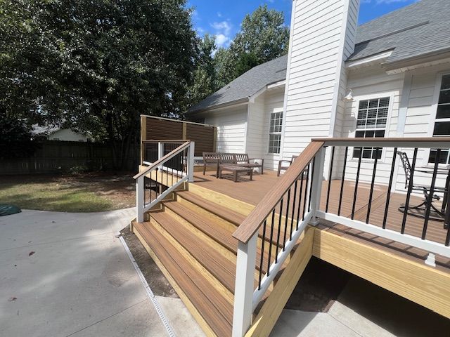 Wooden deck with stairs, built against a white house. Black railing, brown decking, blue sky.