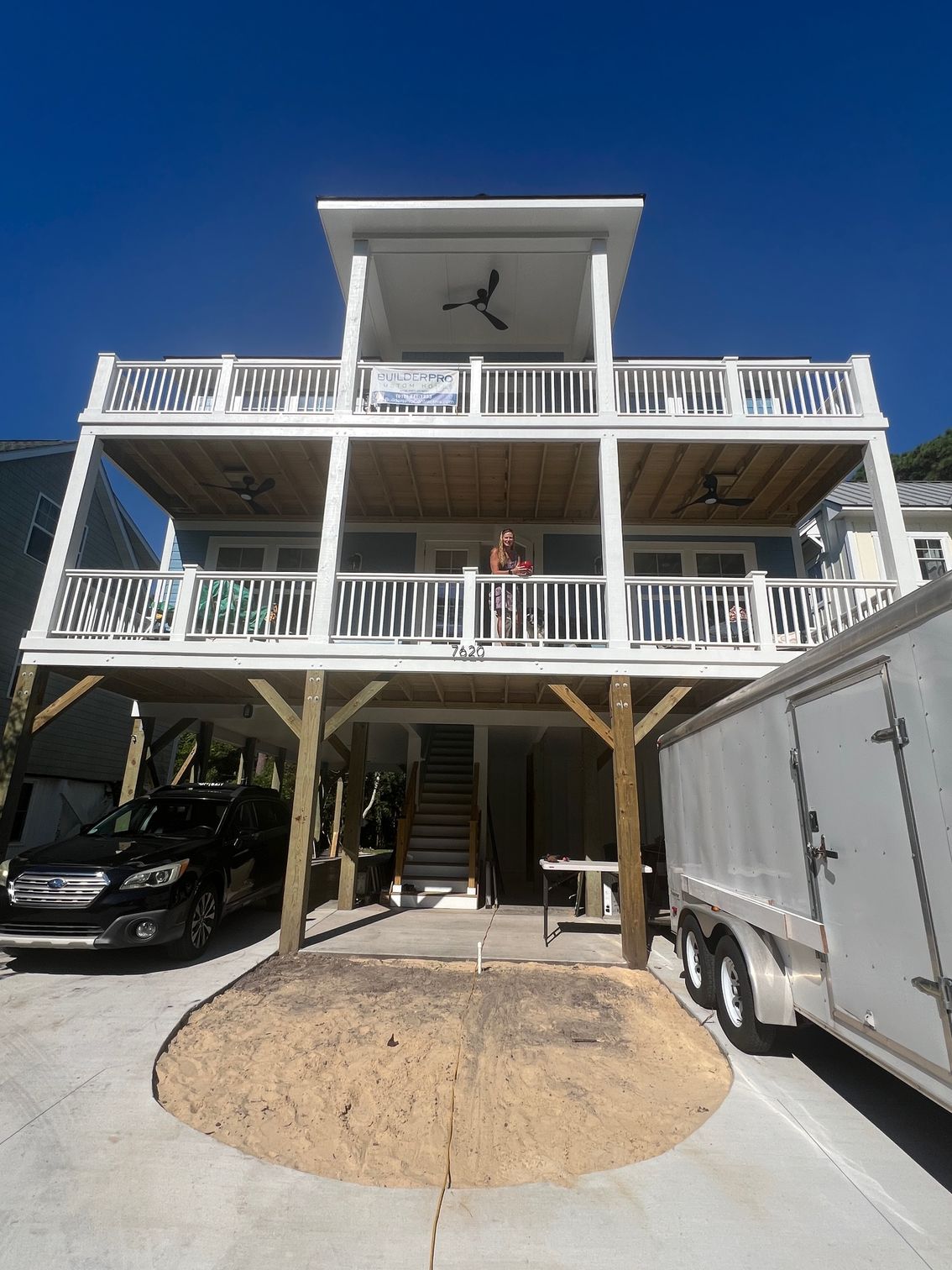 Multi-level house with decks and railings; person on second-story balcony. Trailer and car in front.