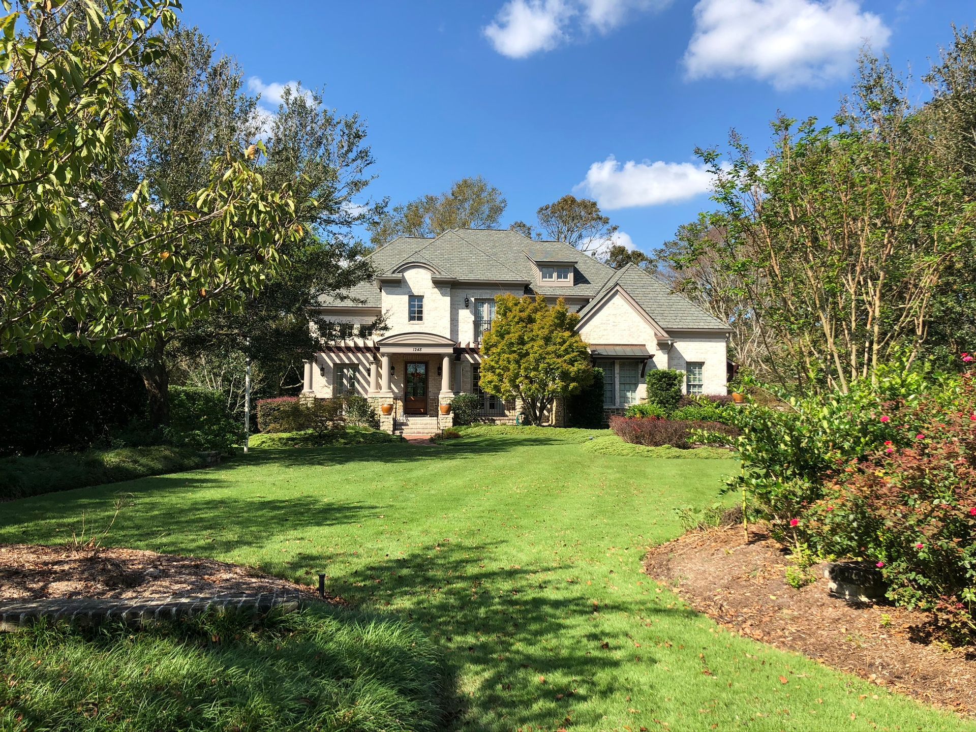 Two-story beige house with a stone facade, green lawn, trees, and blue sky.